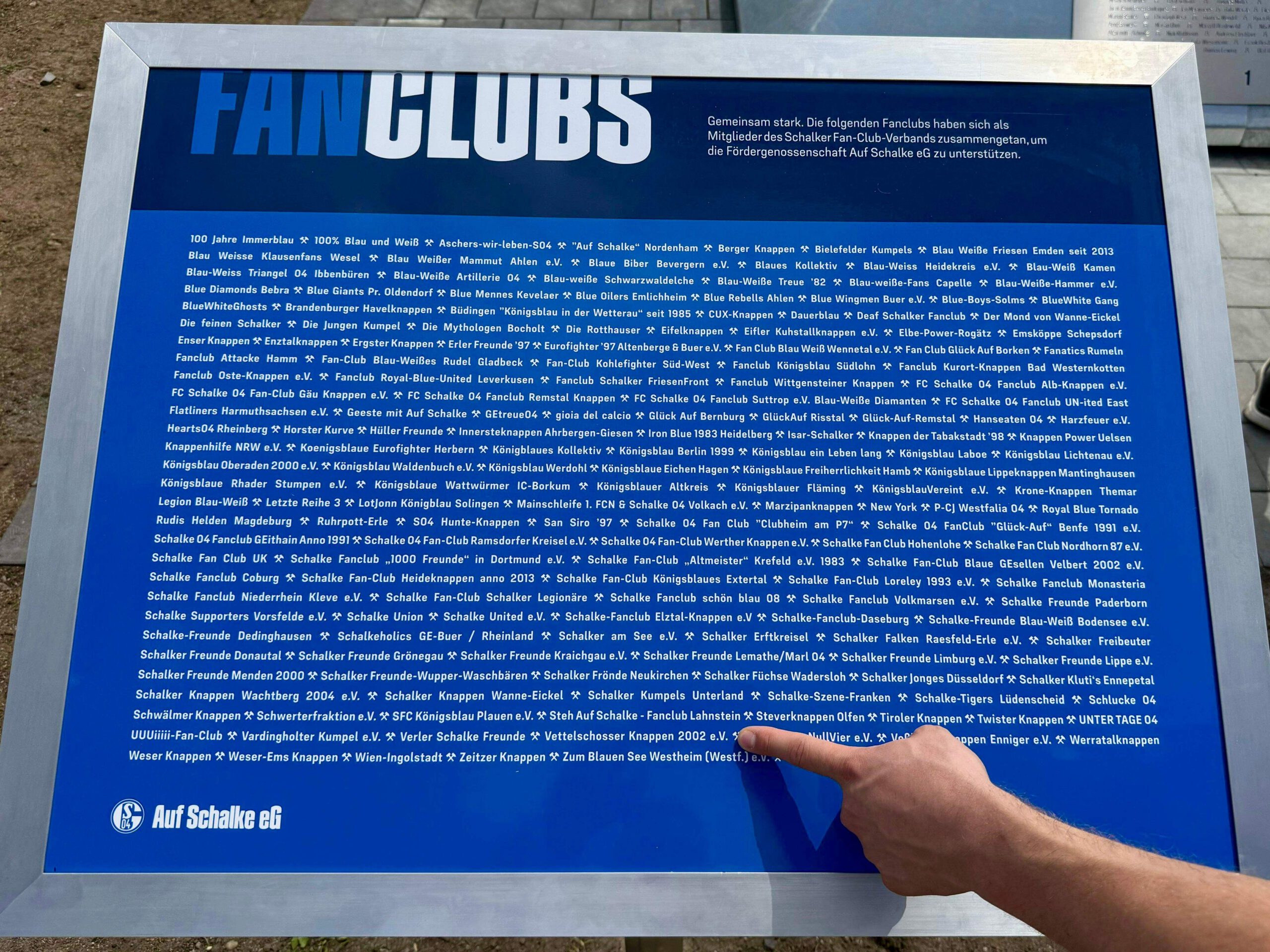 Zu sehen ist eine blaue Schrifttafel mit der Überschrift "Fanclubs". Darunter finden sich sämtliche Fanclubs, die die Genossenschaft Auf Schalke eG unterstützen. Ein Finger zeit auf den Namen der Vettelschosser Knappen 2002 e.V.